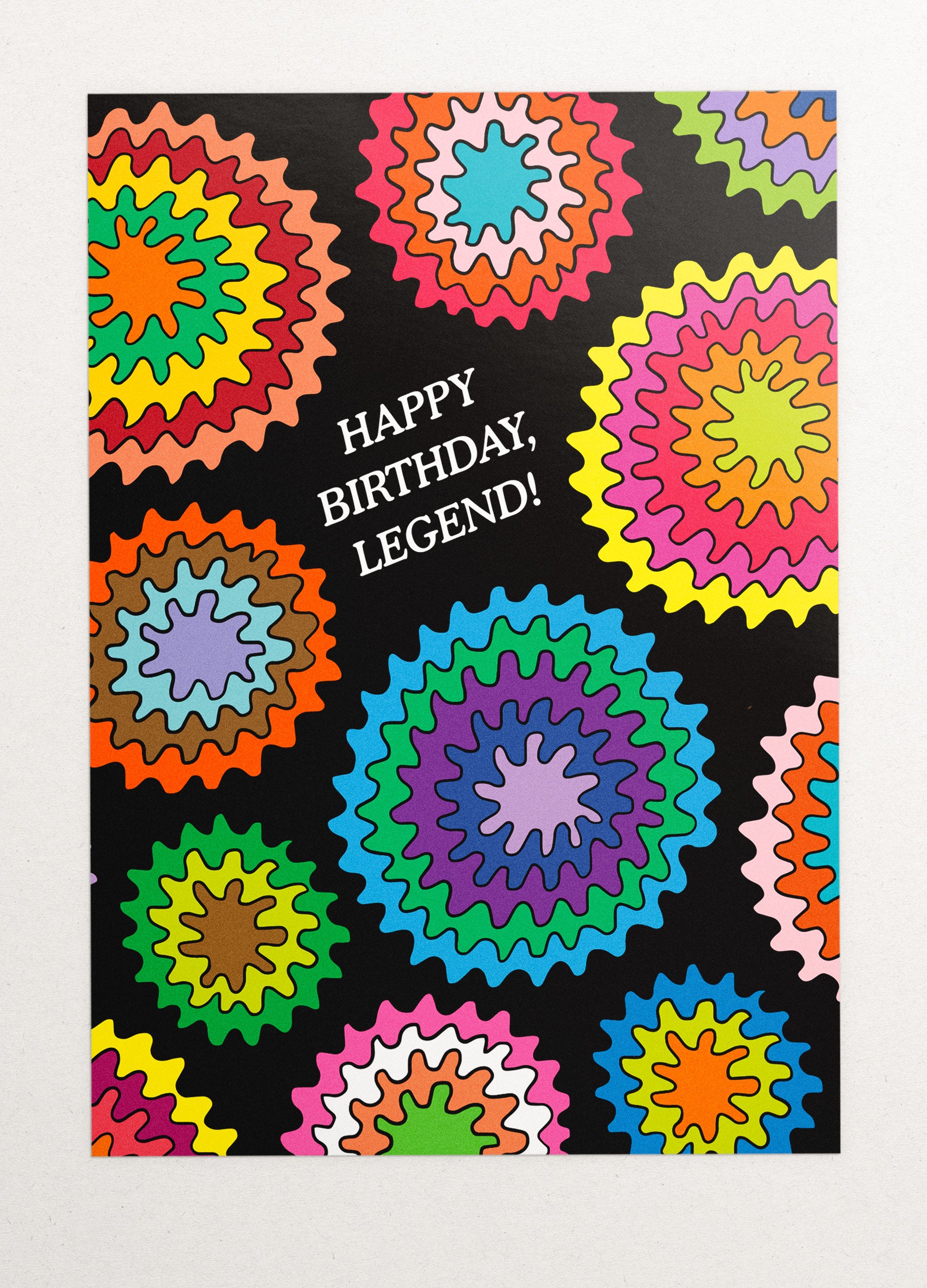 This image is a greeting card by Kiosk that has a black background and a circular wavey burst design. The design has different burst patterns and shapes in pink, orange, green, blue, and purple. The card also has a white text that says “Happy Birthday, Legend!” in a fun and casual font. The card has a playful and cheerful design that conveys a message of celebration and compliments for your birthday. 🎂