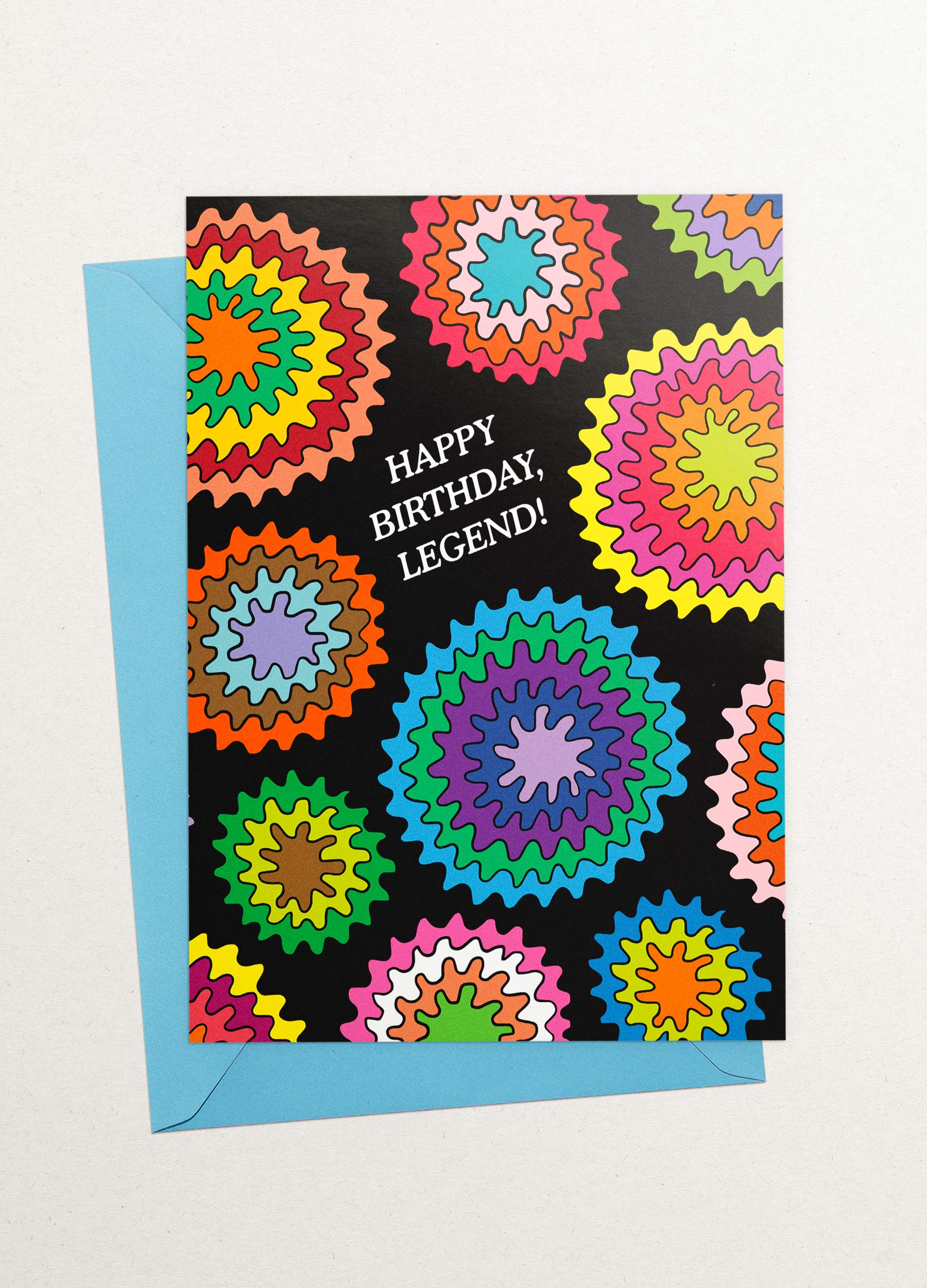 This image is a greeting card by Kiosk that has a black background and a circular wavey burst design. The design has different burst patterns and shapes in pink, orange, green, blue, and purple. The card also has a white text that says “Happy Birthday, Legend!” in a fun and casual font. The card has a playful and cheerful design that conveys a message of celebration and compliments for your birthday. 🎂 Partly showing behind it is a light blue envelope.