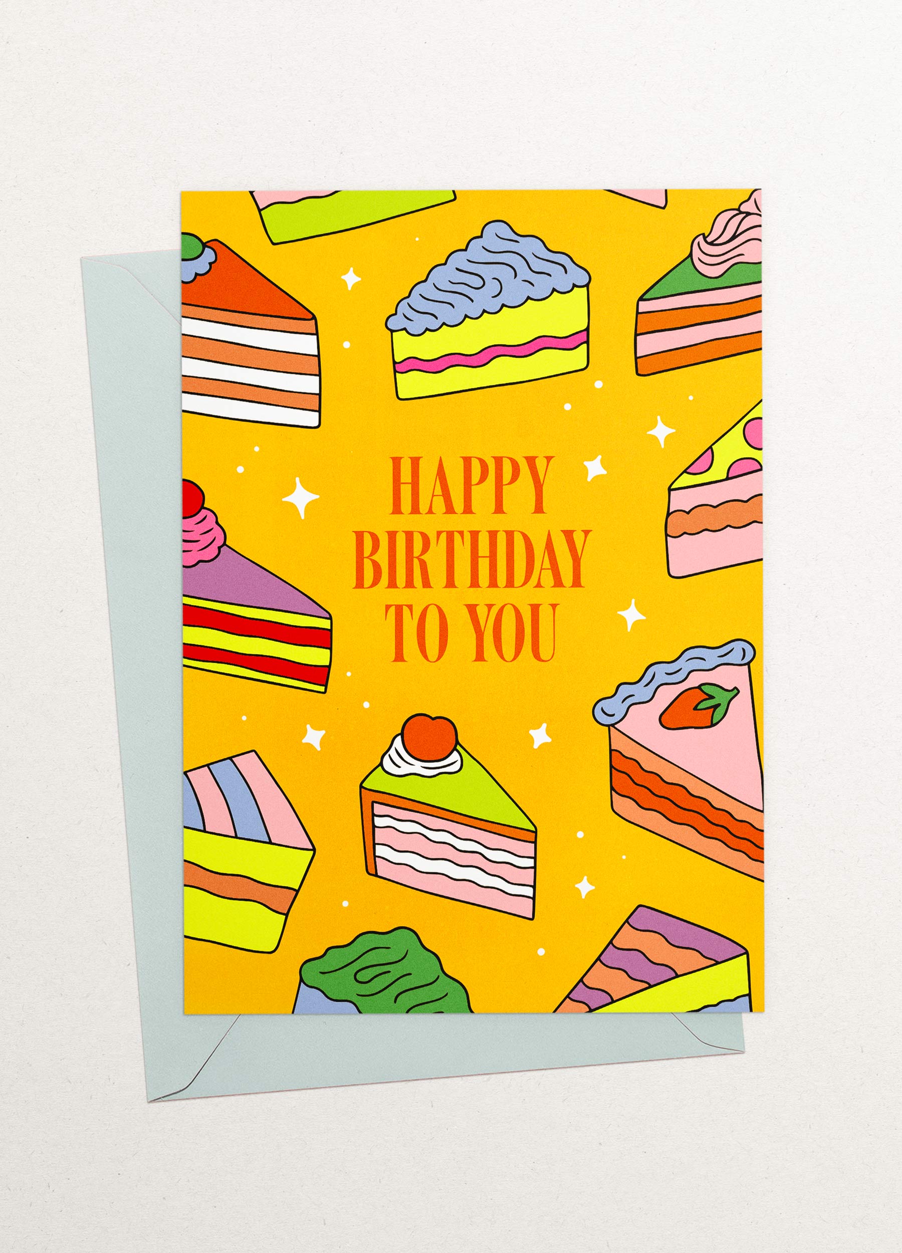 This is an image of a greeting card by Kiosk, that is illustrated by Georgia Perry. The card has a yellow background and illustrations of different types of cake slices scattered across the background. The cakes are in different colors and styles, including pink, green, and orange. The text on the card reads “Happy Birthday to You” in white letters. There are also small white stars scattered across the background. Partially showing behind it is a light blue envelope.