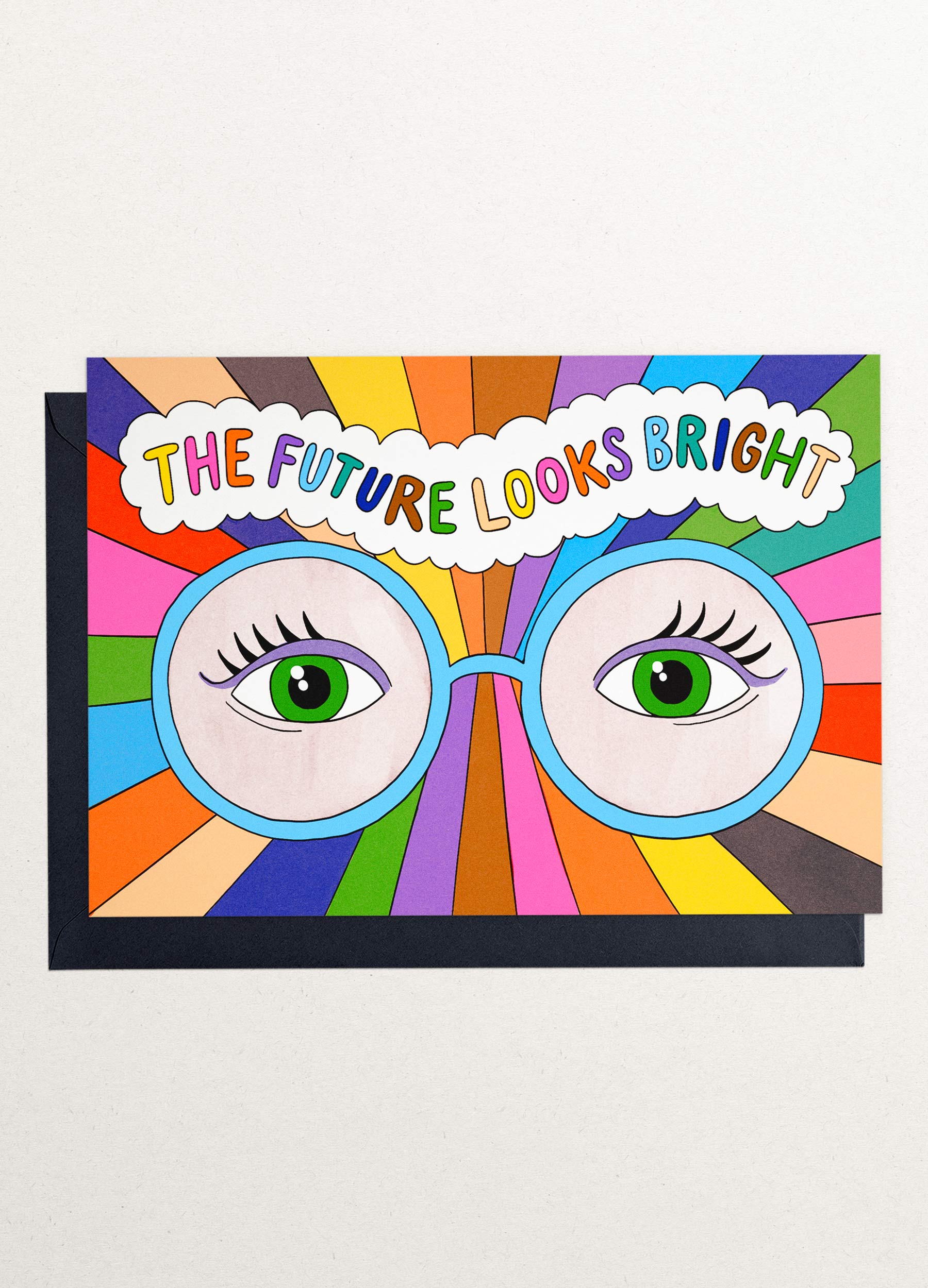 This is an image of a greeting card by Kiosk, that is illustrated by Georgia Perry. The card has an illustration of a pair of green eyes with colorful rays emanating from them. The rays are in various colors such as orange, pink, blue, green, and yellow. Above the eyes, there is a cloud with the text “The future looks bright” written in blue. The background is white and the illustration has a cartoon-like style. Partly showing behind it is a dark navy blue envelope.