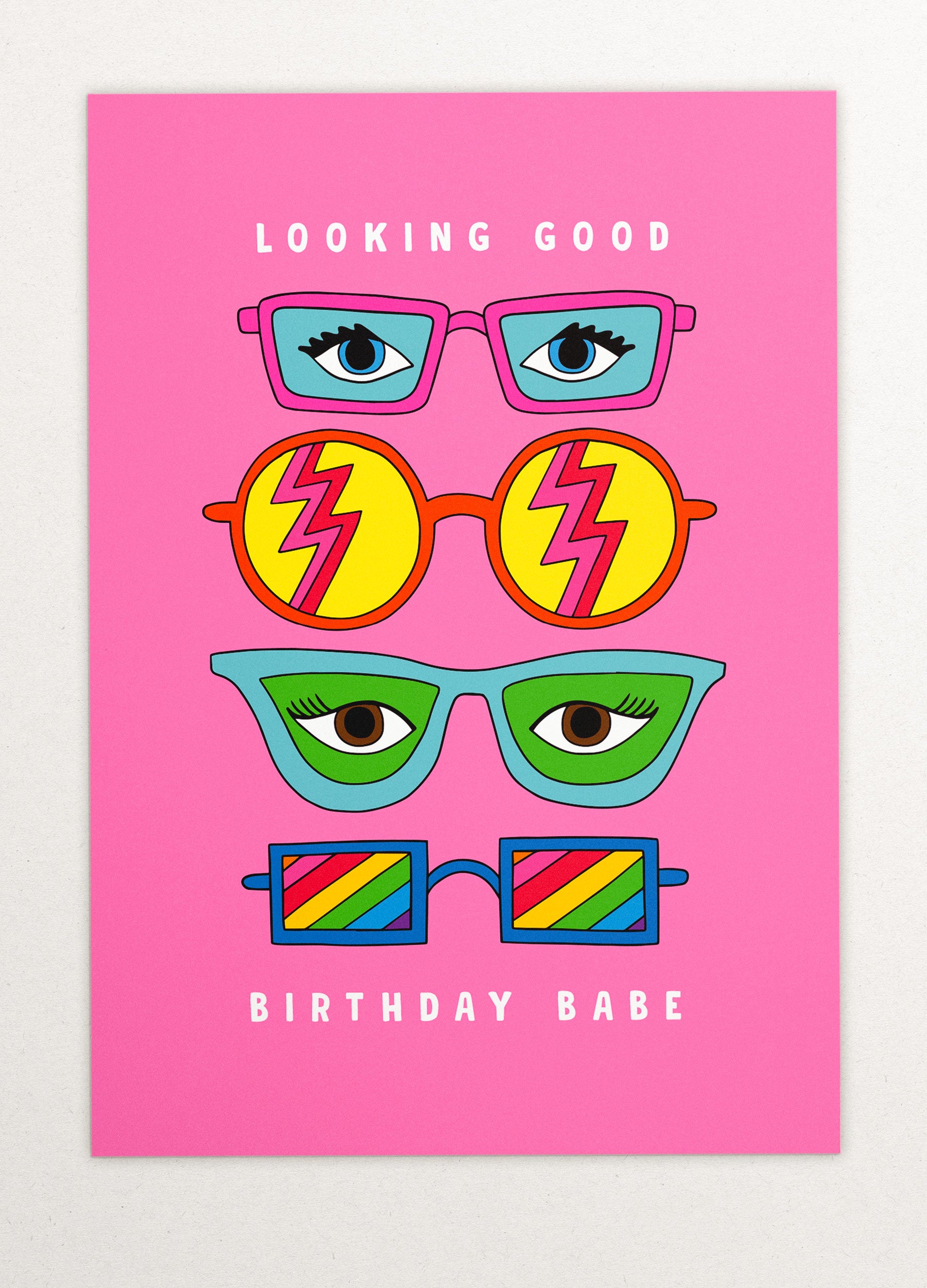 This image is a greeting card by Kiosk that has a pink background. It has three pairs of illustrated glasses on it. The glasses have different shapes, colors, and patterns on the lenses. Above the top pair of glasses is the text “Looking Good”, and below the bottom pair is the text Birthday Babe”. The card has a playful and cheerful design that conveys a message of celebration and compliments for your birthday. 🎂