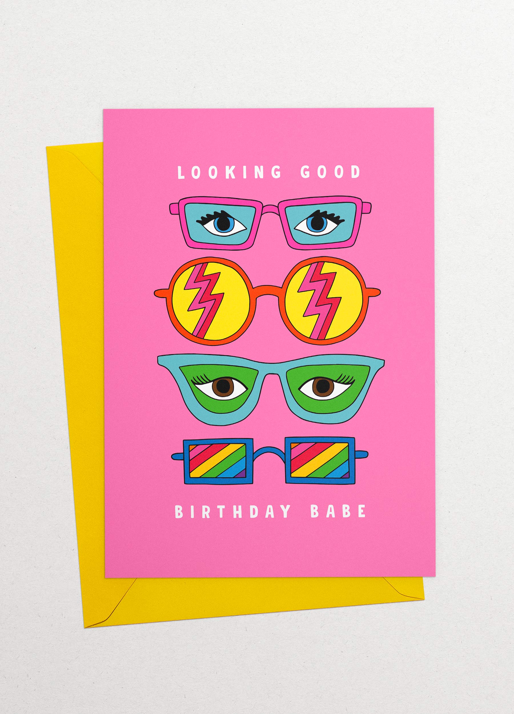 This image is a greeting card by Kiosk that has a pink background. It has three pairs of illustrated glasses on it. The glasses have different shapes, colors, and patterns on the lenses. Above the top pair of glasses is the text “Looking Good”, and below the bottom pair is the text Birthday Babe”. The card has a playful and cheerful design that conveys a message of celebration and compliments for your birthday. 🎂 Partly showing behind it is a yellow envelope.
